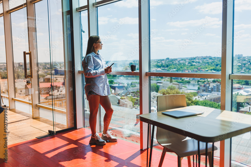 Attractive female blogger with a modern smartphone in her hands stands at the window, thoughtful look and daydreaming, holding a mobile phone, ponders an idea for a publication in social networks