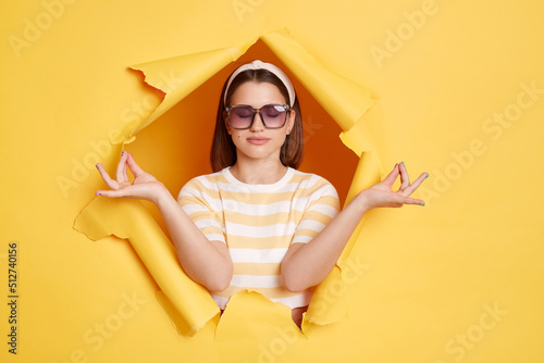 Portrait of relaxed dark haired Caucasian woman wearing striped shirt and hair band posing in yellow paper hole, relaxing, calming down, meditating, keeps eyes closed.