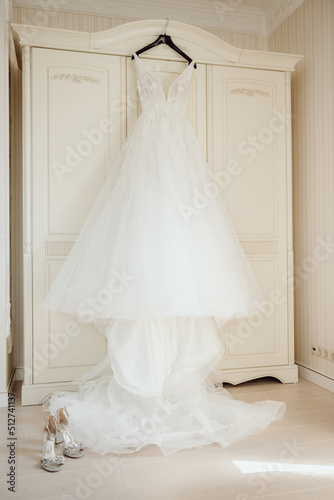 A white beautiful bride's dress hangs on the door of a wooden wardrobe in a bright white room.
