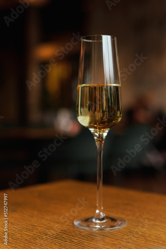 Glass of champagne standing on wooden table in soft focus on naturally blurred dark background. White wine with bubbles. Celebration, happy holidays concept