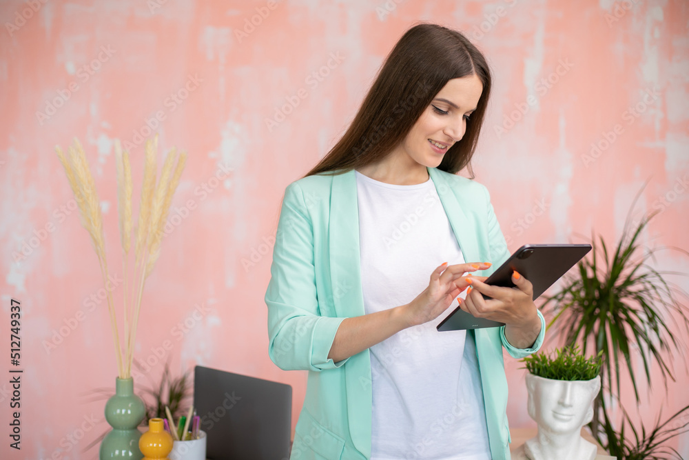 Portrait of cheerful stylish girl in smart casual outfit standing at desk holding tablet in hands Portrait of cheerful stylish girl in smart casual outfit standing at desk holding tablet in hands
