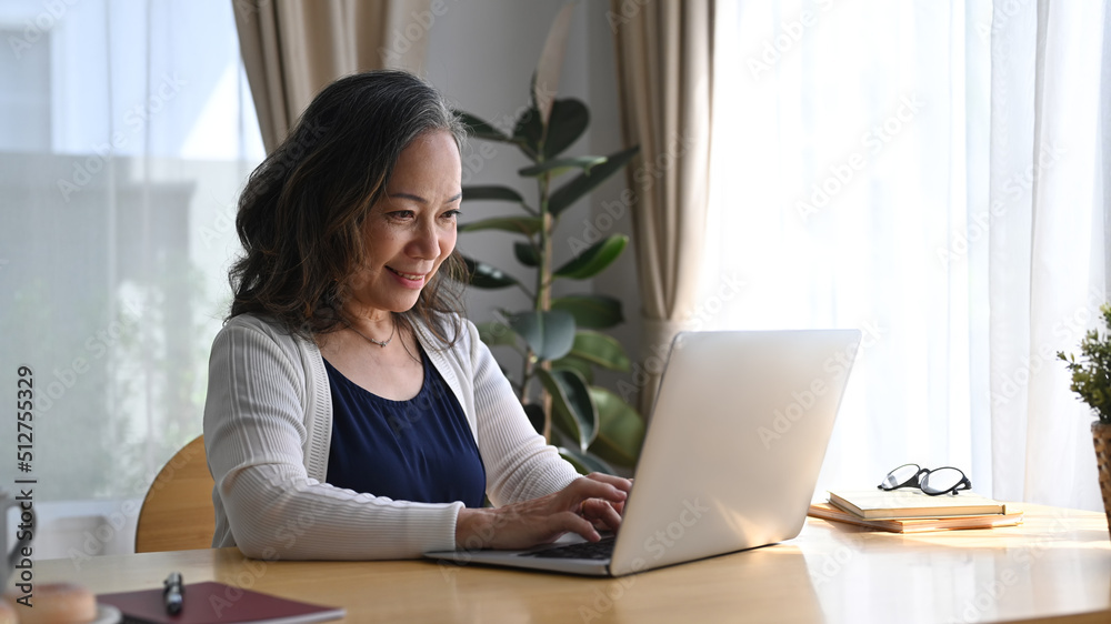 Middle age woman sitting at table in comfortable home and using computer laptop with happy face Middle age woman sitting at table in comfortable home and using computer laptop with happy face
