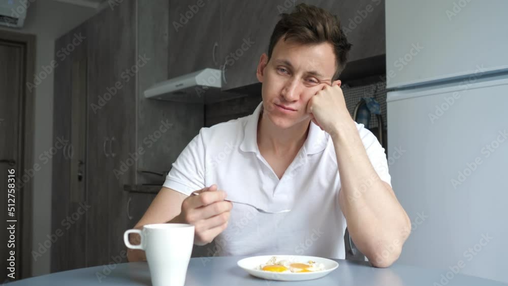 Sleepy man has breakfast and drinks cup of coffee at table. Young father naps and very tired eating in kitchen. Exhausted guy sleeps badly Sleepy man has breakfast and drinks cup of coffee at table. Young father naps and very tired eating in kitchen. Exhausted guy sleeps badly
