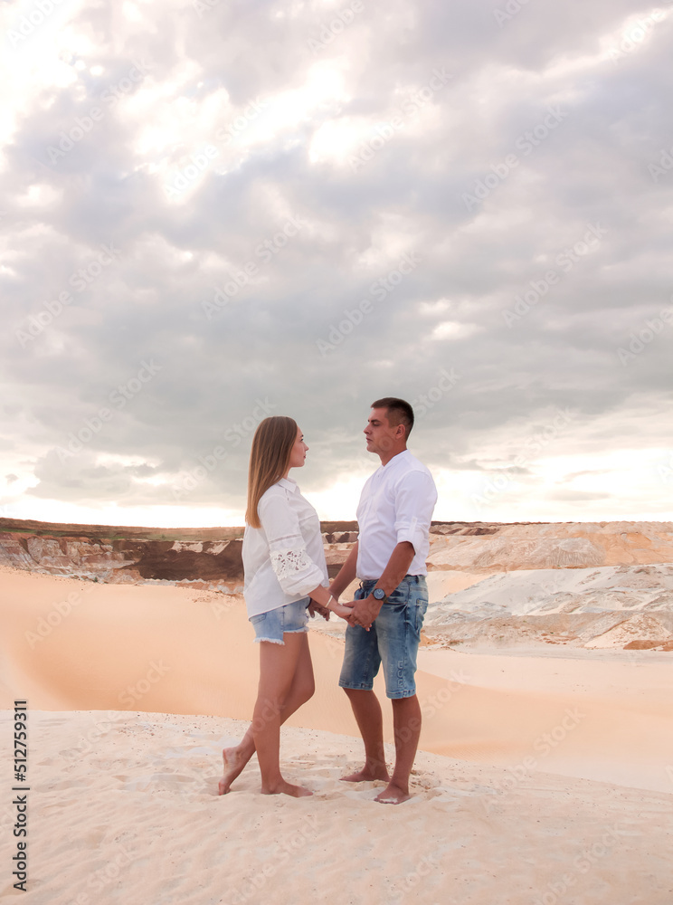 young teen couple in white shirts and jeans are standing each other and holding hands with love on sand career background in desert at sunset. romantic date relationship. lifestyle concept, free space