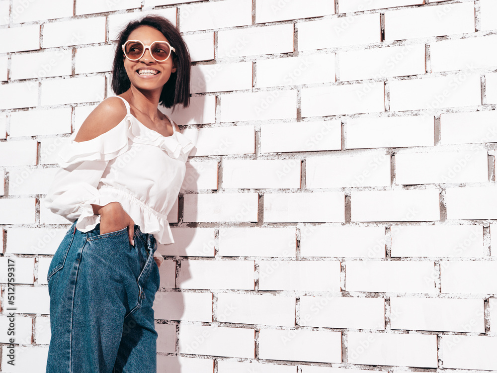 Portrait of young beautiful black woman. Smiling model dressed in summer jeans clothes. Sexy carefree female posing near white brick wall in studio. Tanned and cheerful. In sunglasses Portrait of young beautiful black woman. Smiling model dressed in summer jeans clothes. Sexy carefree female posing near white brick wall in studio. Tanned and cheerful. In sunglasses