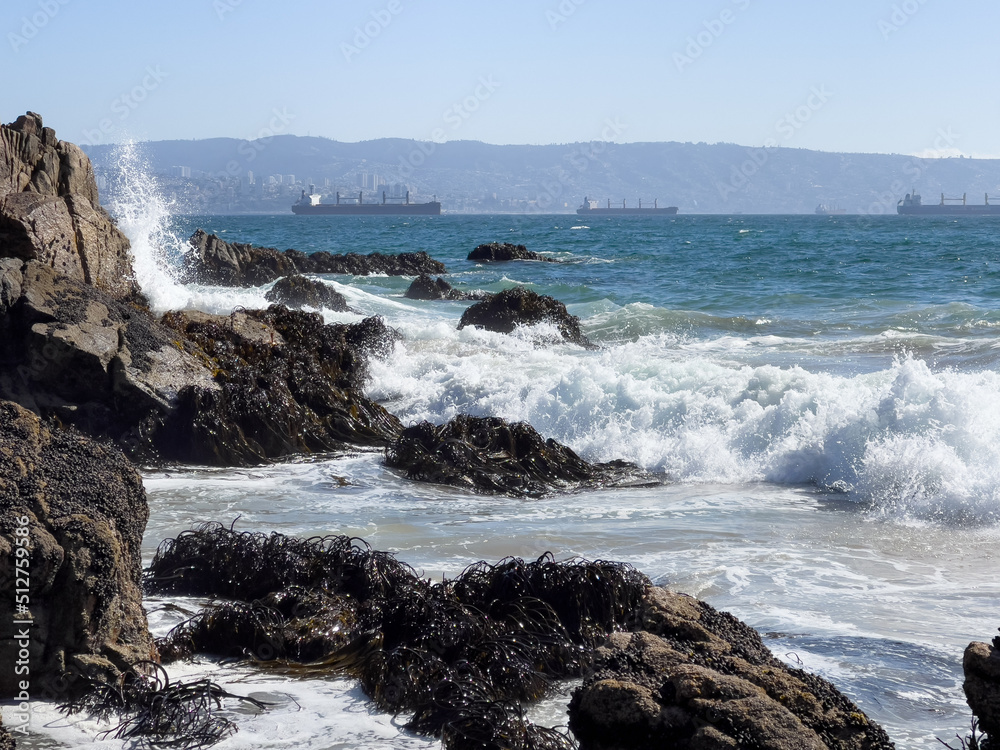 Sea water splash on rocks, algae and cargo ships by Valparaiso harbor on background. Brave waves crashing on shore on sunny day in Chile