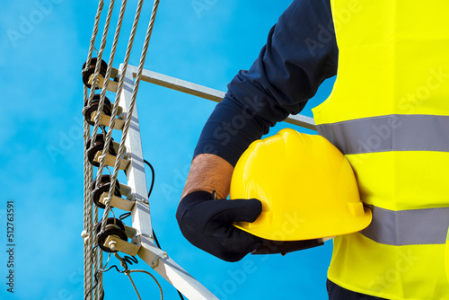 A working electrician in a yellow vest holds a protective helmet in his hand against the background of a power line, stretched wires with insulators.