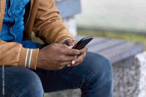 Close-up of the hand of a young black businessman sitting on a bench and working on a smartphone. Fashionable man in a brown jacket uses his phone to chat online with a business partner