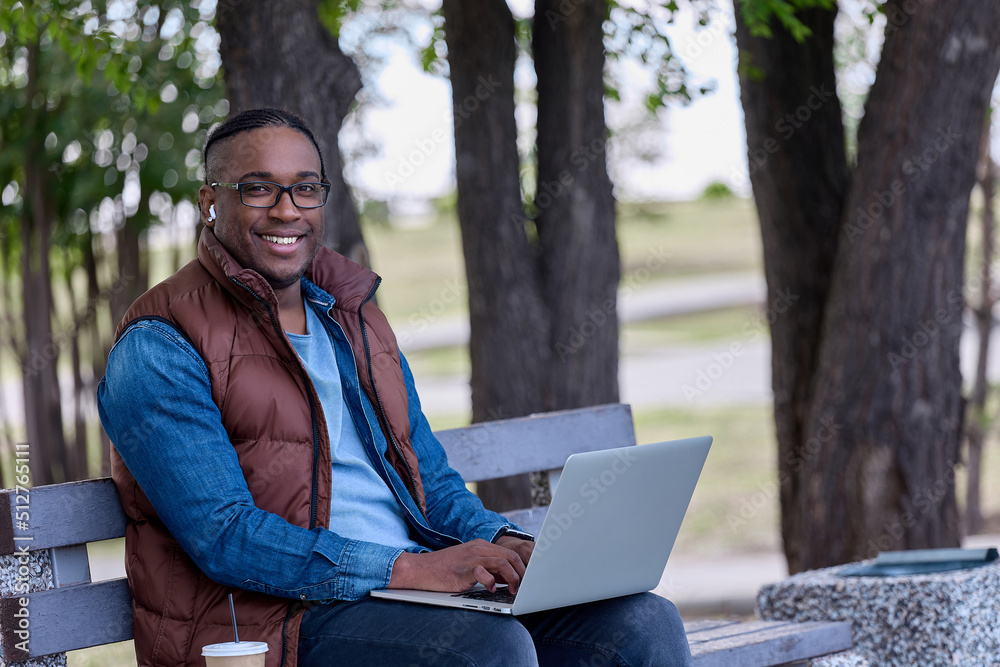 Cheerful man in glasses and a brown vest sits on a bench with a laptop drinking coffee. A popular black blogger reports on the street in a park on a bench in clear weather Cheerful man in glasses and a brown vest sits on a bench with a laptop drinking coffee. A popular black blogger reports on the street in a park on a bench in clear weather