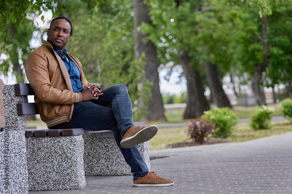 Obraz premium Handsome black man sits on a bench in the park waiting for another person. An African-American man sat down on a bench in an alley in a park, resting from a long road.