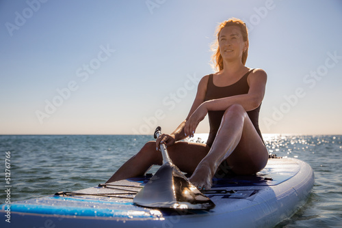 A beautiful young Caucasian slender woman in a black swimsuit sits in a relaxed pose on a sup board in the middle of a calm sea against the blue sky on a summer day. Beautiful sun glare play on the