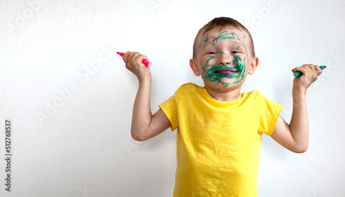 A 4-year-old boy painted his face with a marker. The boy stands against a white wall, holding markers in his hands and smiling. Banner with space for text
