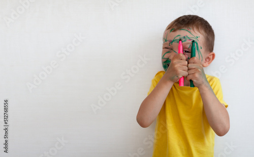 A 4-year-old boy painted his face with a marker. The boy stands against a white wall, holding markers in his hands and smiling. Banner with space for text