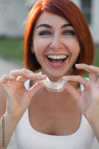 Red-haired Caucasian woman holding transparent mouthguards for bite correction outdoors. A girl with a beautiful snow-white smile uses silicone braces