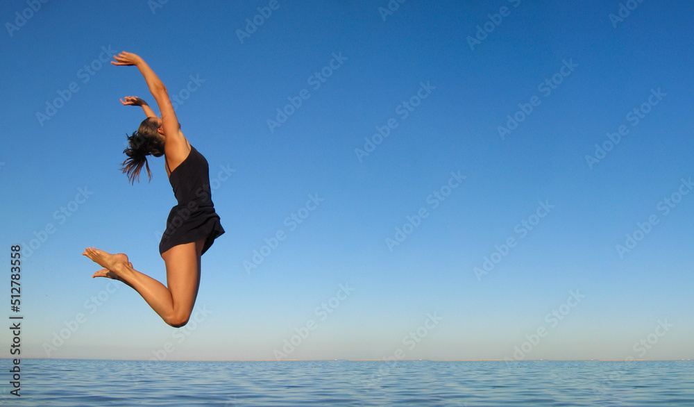 Concept of happiness, freedom, vacation, relaxation. Young girl jumps high in air over sea against sky in dark blue short dress Concept of happiness, freedom, vacation, relaxation. Young girl jumps high in air over sea against sky in dark blue short dress