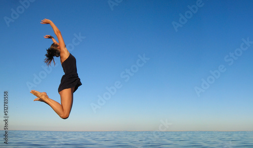 Concept of happiness, freedom, vacation, relaxation. Young girl jumps high in air over sea against sky in dark blue short dress