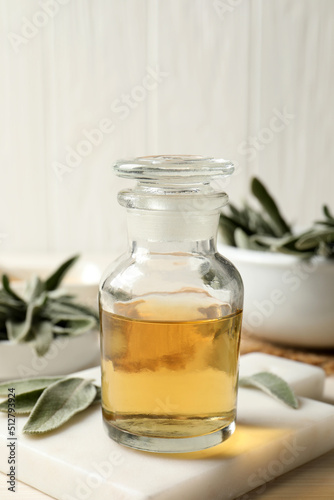 Bottle of essential sage oil and leaves on white wooden table, closeup