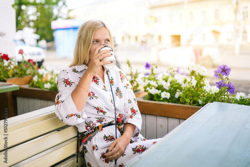 Girl in a white dress is drinking coffee. Beautiful blonde with a mug on the veranda with flowers. People in the urban environment