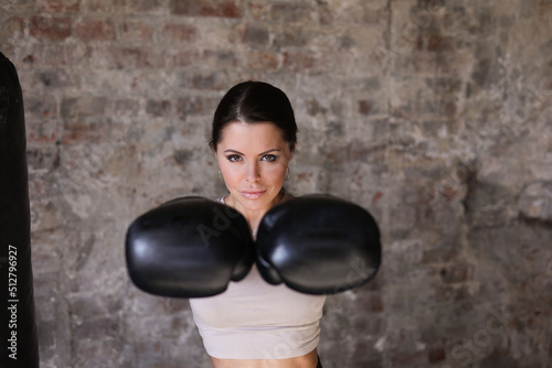 Beautiful middle-aged thin sporty woman boxing and bitting punching bag. Strong muscular young girl practicing box in black boxing gloves in gym. Fit slim woman in gris gym in black pants © Tatyana