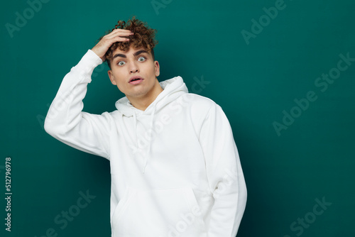 a cute  funny  emotional man stands on a green background in a white hoodie and looks at the camera with his mouth wide open  holding his face with his hands