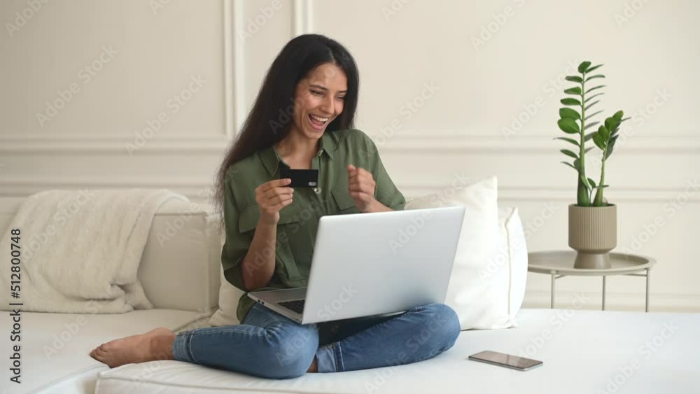 Convenience to do shopping without leaving home. Happy Indian woman holds debit card sitting on the couch at home with a laptop on laps, making purchases, ordering food, enjoying buying online Convenience to do shopping without leaving home. Happy Indian woman holds debit card sitting on the couch at home with a laptop on laps, making purchases, ordering food, enjoying buying online