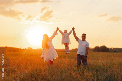 Parents play and lift up by the hands their daughter on meadow at sunset.
