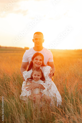 Young family with daughter sits on meadow among grass at sunset.