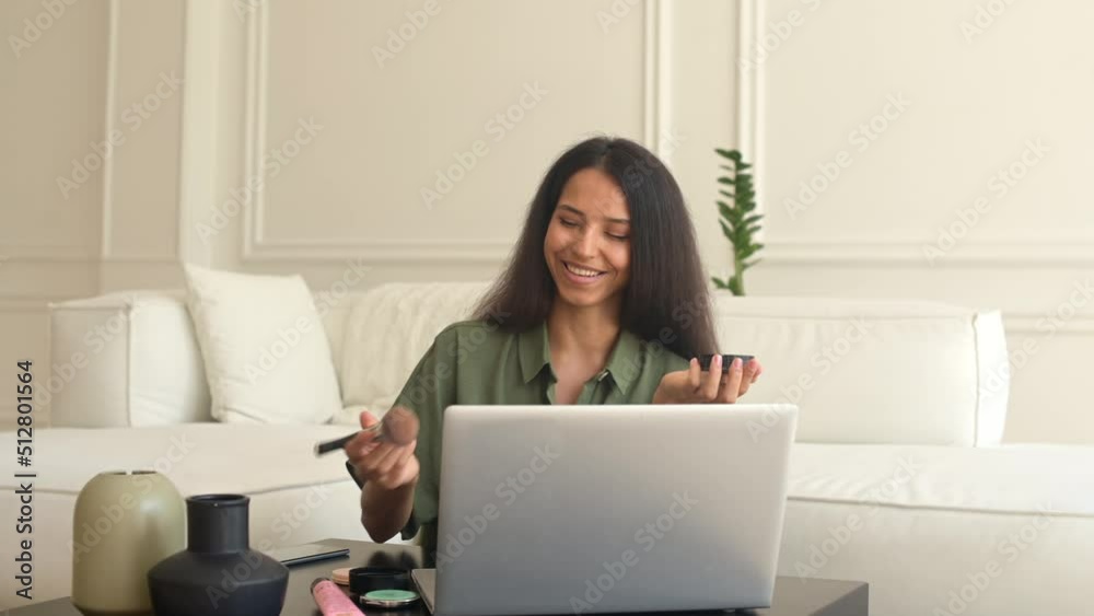 Smiling indian woman applying powder with brush and giving makeup tutorials online, sits in front of the laptop, smiling eastern girl teaching to do makeup by self, records tutorial. Hobby concept Smiling indian woman applying powder with brush and giving makeup tutorials online, sits in front of the laptop, smiling eastern girl teaching to do makeup by self, records tutorial. Hobby concept