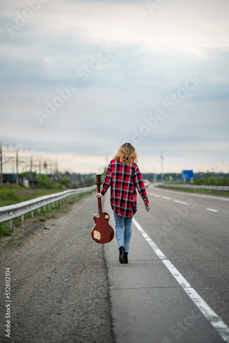 Woman with curly hair, in an unbuttoned plaid shirt, jeans, high heels, walking along the highway towards the horizon (with back to the viewer) with electric guitar. Hitchhiker or traveler musician