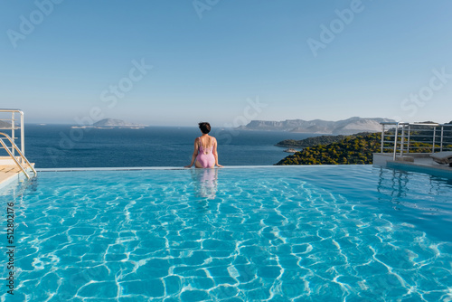 Beautiful young woman swimming in pool and enjoying the epic view over the sea during her summer vacation time.The end of the world. Luxury lifestyle photo of young woman on summer vacation