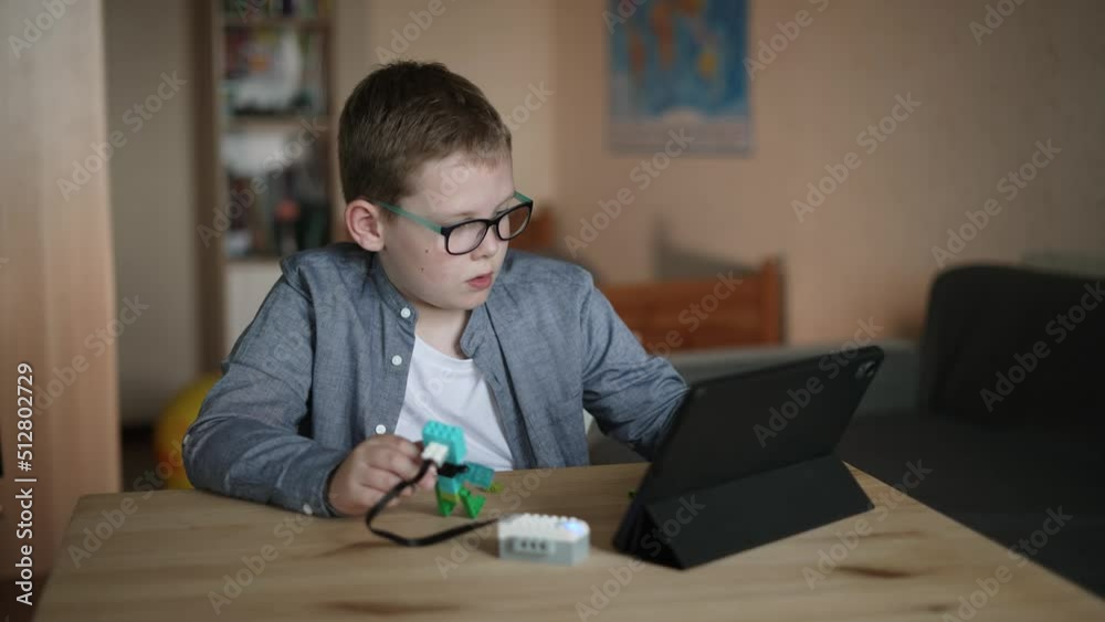 Caucasian boy in glasses sits at table, tries to program movement algorithm of toy from constructor on tablet. Child is surprised, embarrassed because of failed attempt, looks at screen in confusion. Caucasian boy in glasses sits at table, tries to program movement algorithm of toy from constructor on tablet. Child is surprised, embarrassed because of failed attempt, looks at screen in confusion.