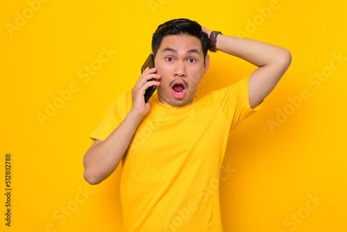 Shocked young Asian man in casual t-shirt talking on mobile phone and touching head isolated on yellow background. People lifestyle concept