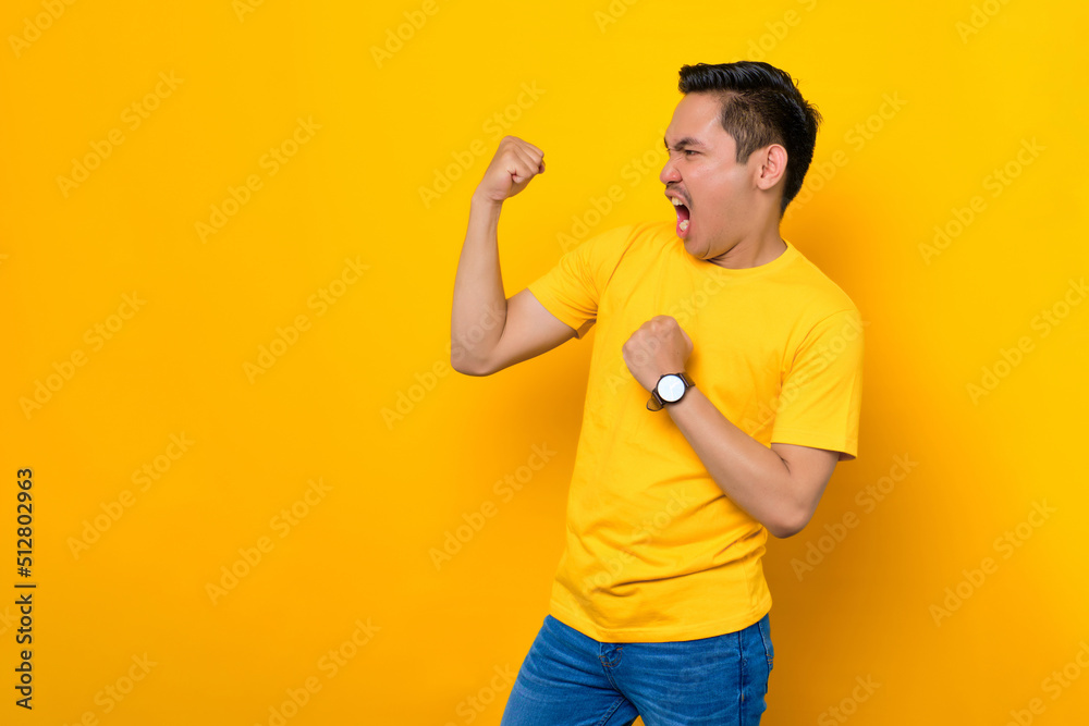 Excited young Asian man in casual t-shirt celebrating success with raised fist isolated on yellow background. People lifestyle concept Excited young Asian man in casual t-shirt celebrating success with raised fist isolated on yellow background. People lifestyle concept