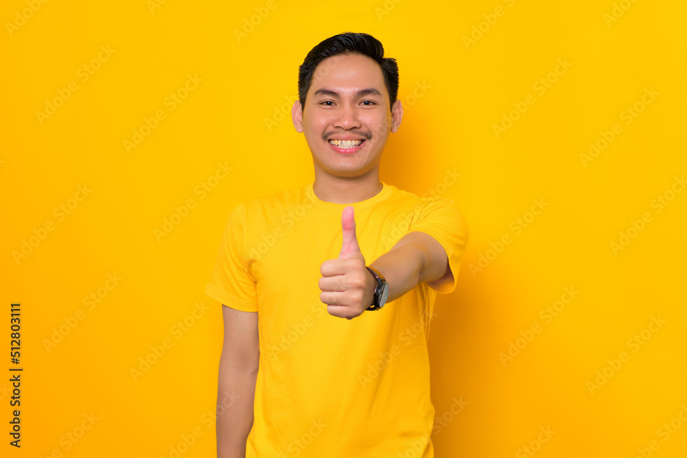 Smiling young Asian man in casual t-shirt showing thumb up and gesturing sign of approval isolated on yellow background. People lifestyle concept Smiling young Asian man in casual t-shirt showing thumb up and gesturing sign of approval isolated on yellow background. People lifestyle concept