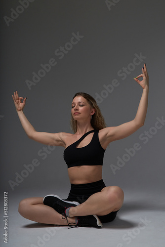young fit sporty attractive blond caucasian woman in black sportswear and sneakers practicing lotus asana on grey background. yoga pose, sport, fitness training, healthy lifestyle, workout