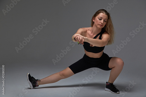 young fit sporty attractive blond caucasian woman in sportive bra and leggings does stretching exercise for legs at grey background. sport, fitness training, healthy lifestyle, workout