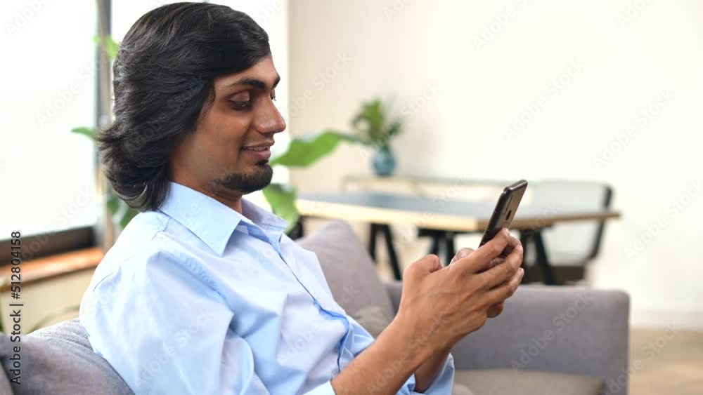 Serene young indian man holds smartphone sitting on the couch at home, eastern guy chatting online with friends, spend leisure time in social media, texting, scrolling news feed, shopping online Serene young indian man holds smartphone sitting on the couch at home, eastern guy chatting online with friends, spend leisure time in social media, texting, scrolling news feed, shopping online