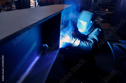 A welder in protective overalls and a welding mask welds metal structures by welding in a hard-to-reach place.
