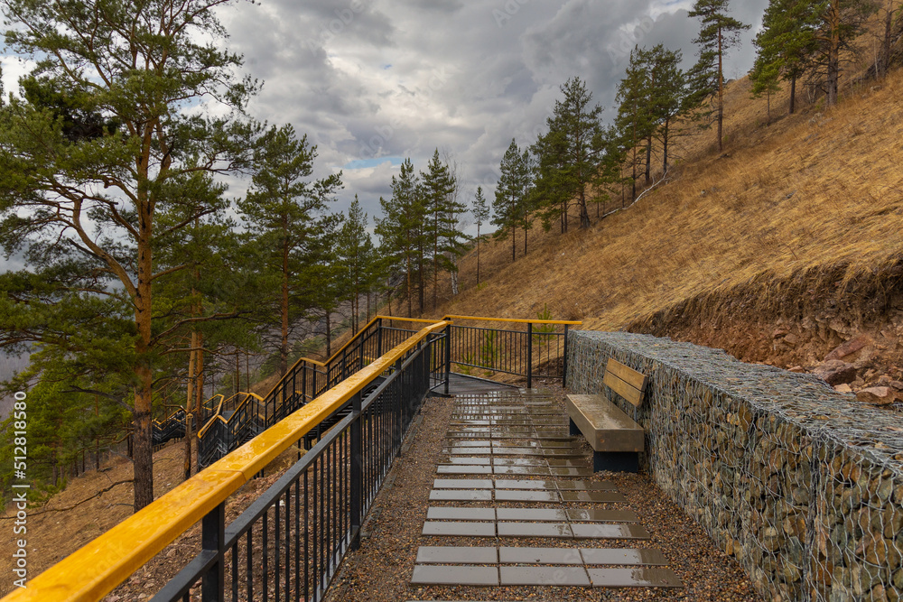 Empty tourist trail in rainy springtime day in the mountain hillside. Metal staircase with wooden railing among pine trees on Torgashinsky ridge in Krasnoyarsk, Russia