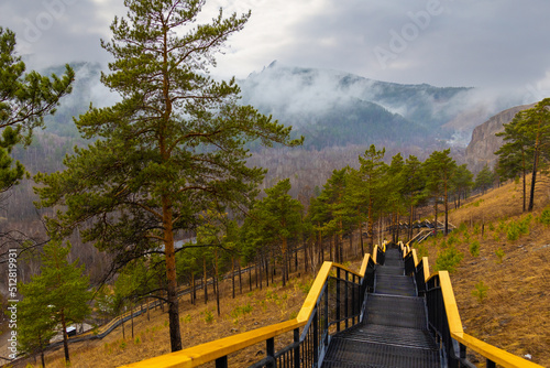 Tourist staircase in rainy springtime day on Torgashinsky ridge in Krasnoyarsk, Russia. Empty metal steps with wooden railing among pine trees in the hillside