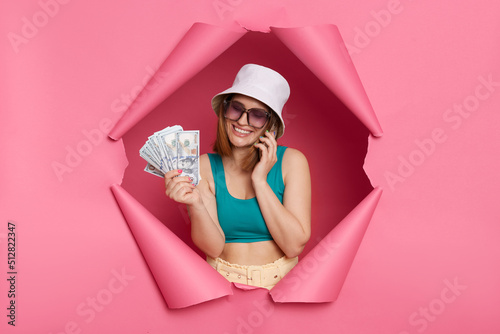 Image of smiling positive Caucasian woman wearing tank top and panama posing in torn pink paper wall, talking phone and holding dollar banknotes, boasting big salary.