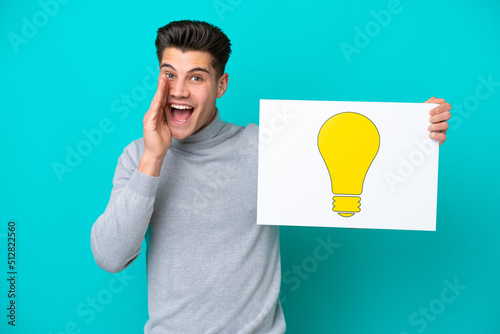 Young handsome caucasian man isolated on blue bakcground holding a placard with bulb icon and shouting