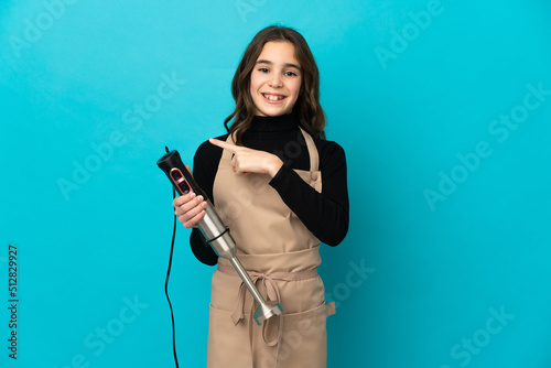 Little girl using hand blender isolated on blue background pointing to the side to present a product