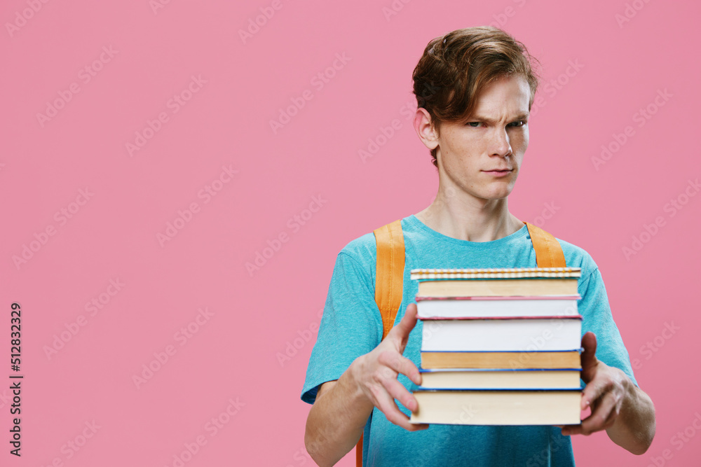 funny, red-haired student in a blue T-shirt and with an orange backpack on his back, holds a heavy stack of books in his hands and looks at the camera with surprise. Horizontal photo with empty space funny, red-haired student in a blue T-shirt and with an orange backpack on his back, holds a heavy stack of books in his hands and looks at the camera with surprise. Horizontal photo with empty space