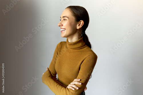 Laughing youthful girl of 20s with perfect sense of humor in stylish turtleneck standing over white background with crossed arms, laughing at funny joke, enjoying leisure time. People and emotions