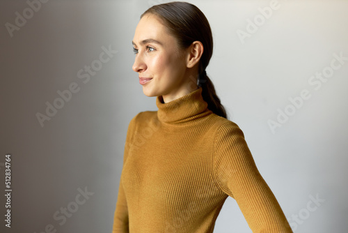 Side view portrait of lovely inspired young attractive Caucasian woman looking aside with shy embarrassed smile posing at camera in turtleneck, isolated on white studio background. Positive emotions