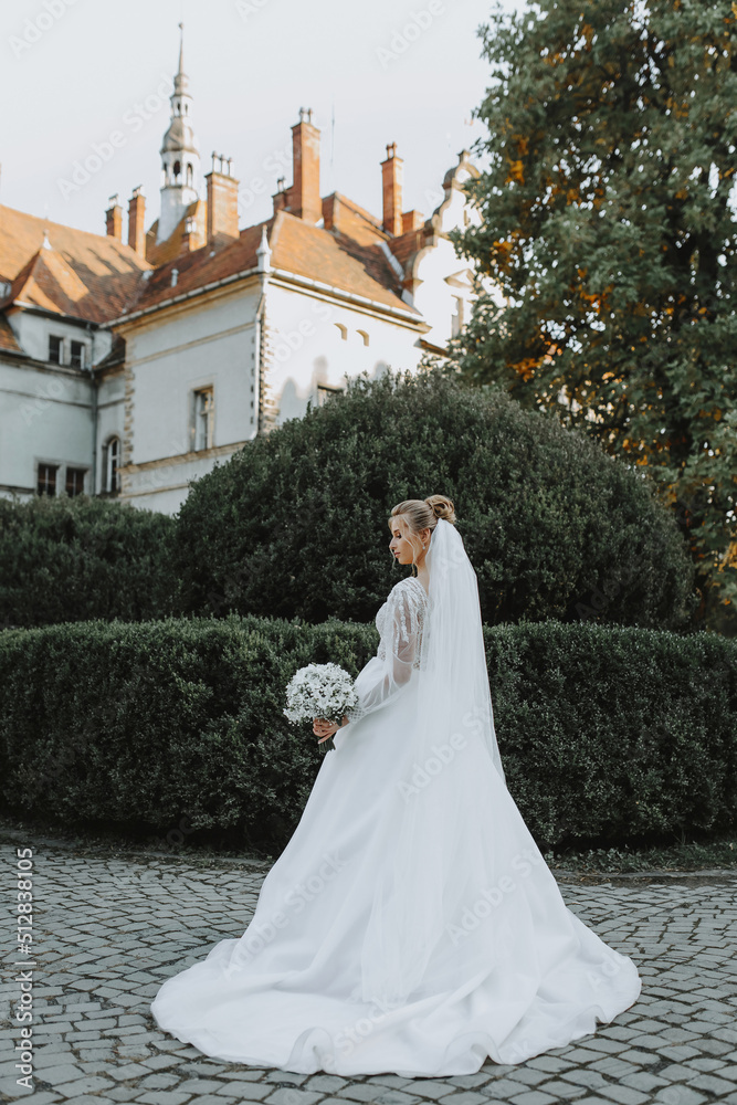 The bride in a beautiful white dress with a bouquet of white flowers stands in the garden against the backdrop of a beautiful old castle.