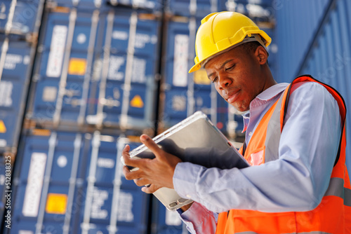 the logistic worker is wearing a yellow safety helmet and holding the smart tablet in a Cargo freight ship for import export container yard with blue sky and airplane background. logistic manager.