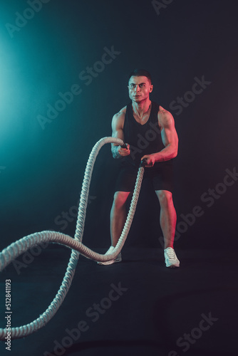 Athletic looking bodybulder work out with battle rope on dark studio background with smoke. Strength and motivation