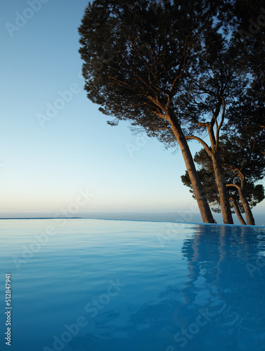 Infinity edge pool with trees and blue sky in background. Beautiful outdoor swimming pool at resort or luxury holiday villa. Romantic getaway with sea view for travelling in summertime © Dhoxax/peopleimages.com